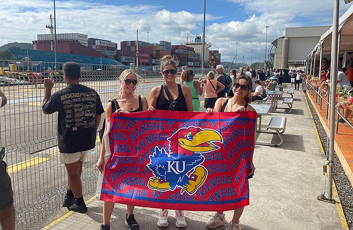 Three girls hold a red and blue flag with the KU Jayhawk and "Kansas" written in a pattern. Shipping containers on a boat stands in the background.