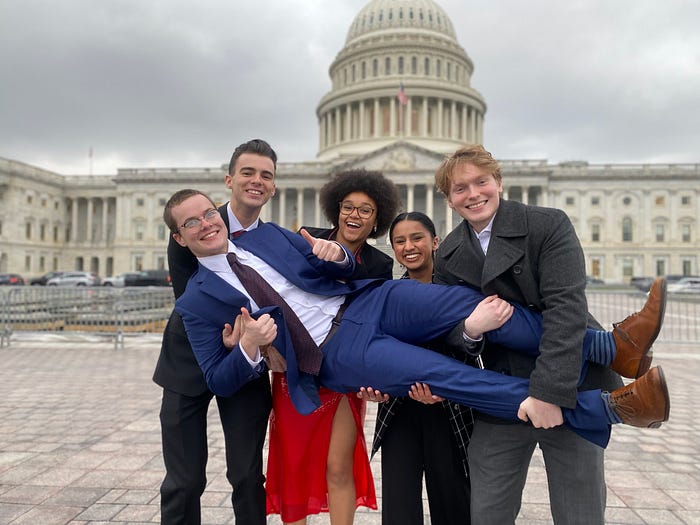 A group of students wearing professional clothing stand in front of the capital building. 