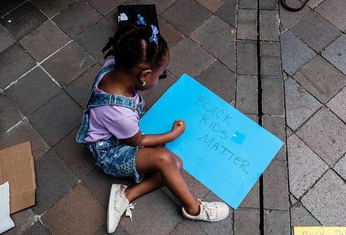 A young Black girl sits on the ground with a bright blue sign that says “Black Kids Matter.”