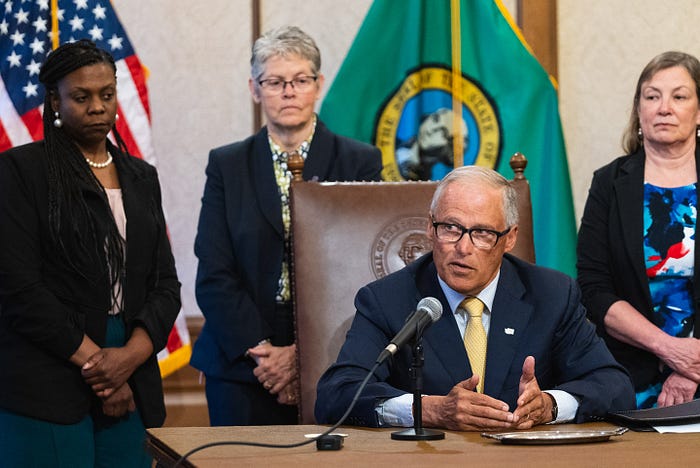 Gov. Jay Inslee is seated and speaking from a wooden table while surrounded by lawmakers.