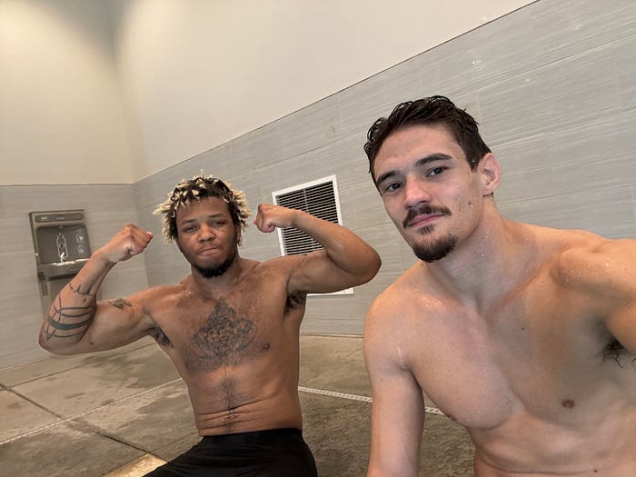 Two men, one flexing, smile at the camera sitting outside of a pool