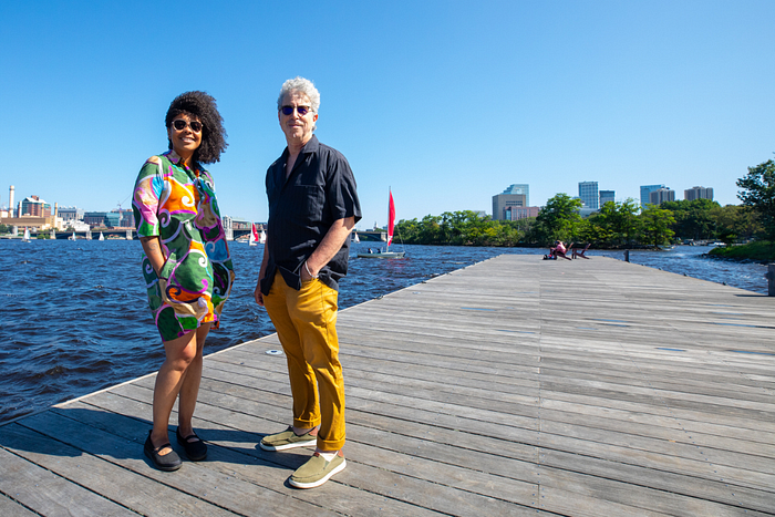 a woman in a vivid printed dress and a man in a blue shirt and gold trousers stand on a dock in the sunshine