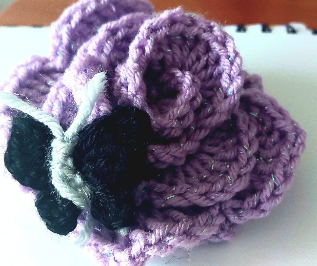 Crochet work coiled into the form of a rose. A crocheted butterfly sits at the side of it.