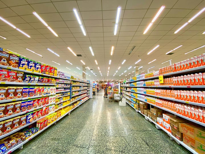 A person stands in a brightly lit supermarket aisle, looking overwhelmed by the sheer number of products on the shelves.