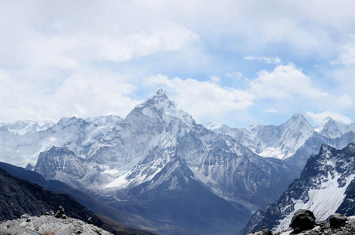 A lone hiker stands on a high mountain ridge, looking out over a breathtaking panorama of clouds and snow-capped peaks.