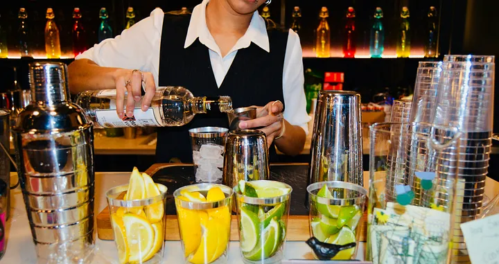 Bartender pouring a drink surrounded by glasses, garnishes and other bar fixtures.