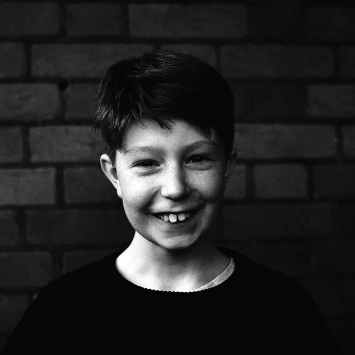 A grayscale image of a boy wearing a dark sweater standing in front of a brick wall.