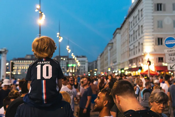 A French street filled with people on a summer’s evening. A young child wearing a France football shirt sits on a parent’s shoulders