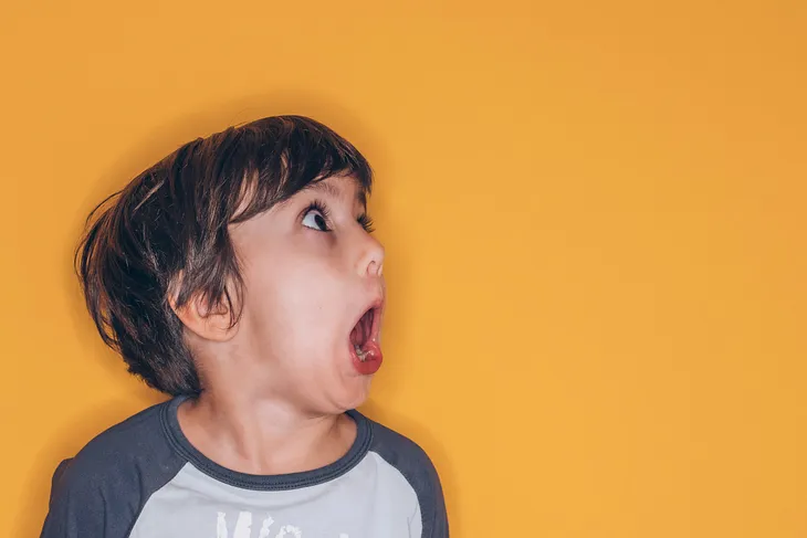 Photograph of a young White child in profile, on the left of the image, with their mouth and eyes wide open, as though in shock. The background is bright yellow.