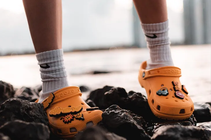 Photo of a child’s feet standing on a rocky terrain near water. The feet have long white Nike socks and colorful orange Crocs.