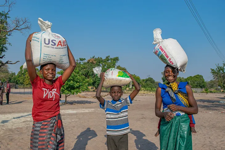 At left, a young Zambian woman carries a bag of grain labeled “USAID.” In the middle, a young boy carries a bag of food atop his head. At right, a smiling Zambian woman carries a bag of food atop her head, with a baby in a sling on her back.