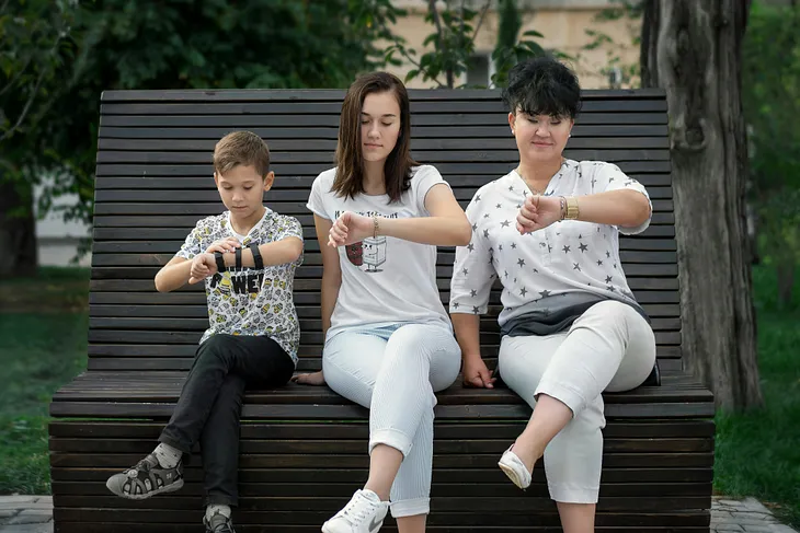 A child, a young woman and a middle-aged woman sit on a bench, looking at their clocks