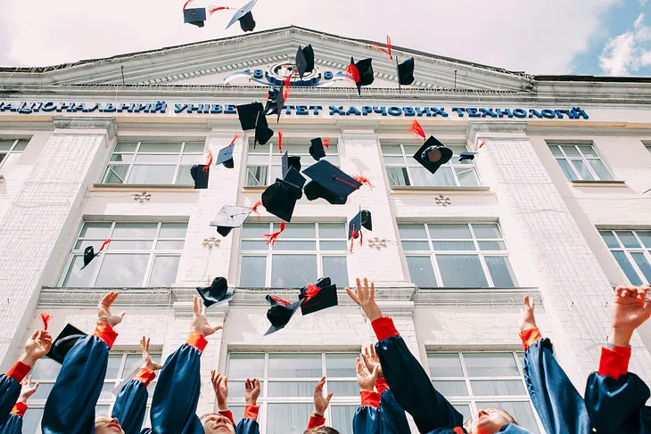 A picture showcasing the hands of many graduates throwing their navy blue graduation hats up in the air. The tassel is red. Their sleeves are blue with red at their wrist. Though there are many hands, only 3 students’ face is shown watching their hats up in the air. There is a building in the background with nine windows; each row has three horizontal windows. 19 hats are all thrown up in the air at different heights.