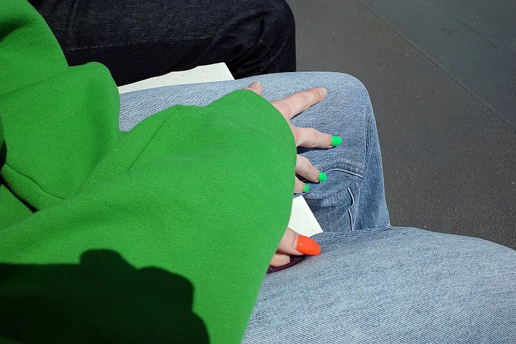 Hands folded on a lap, one hand has green painted nails and the other hand has orange