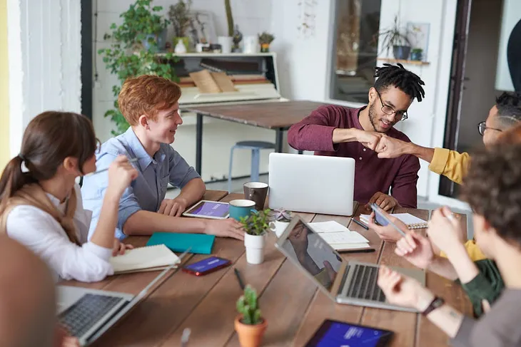 A happy team of young professionals in casual dress working in an office and having a meeting around a table, with two people fist bumping