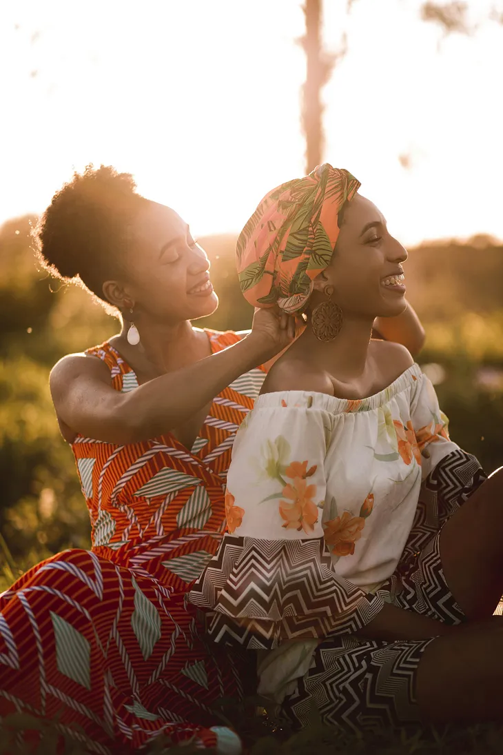 Two beautiful ladies wearing colorful afro-centric clothing are standing together outside in the backdrop of green woods.