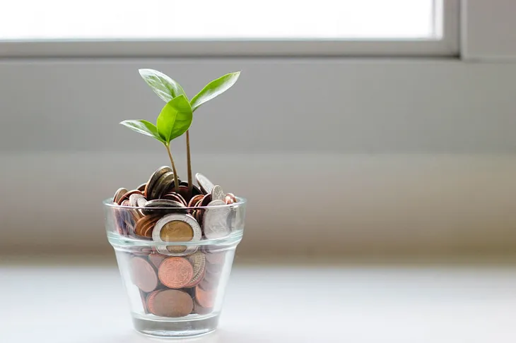 A pile of coins in a glass jor with green shoots growing out of it.