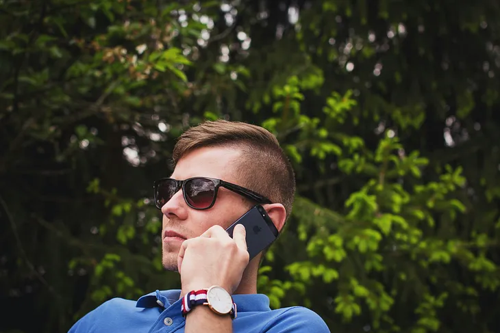Red-headed man wearing sunglasses is listening to his phone in front of a tree.