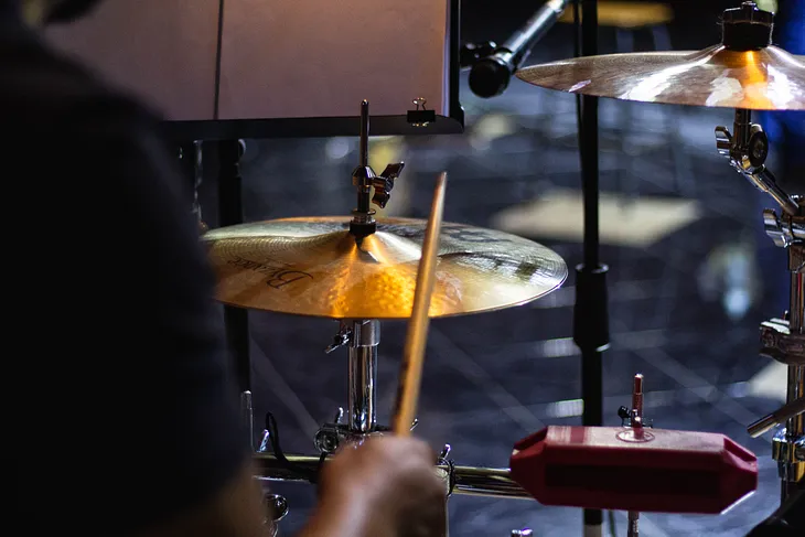 Drummer’s right hand on cymbal, up close, with cymbals, microphone and music stand in background.