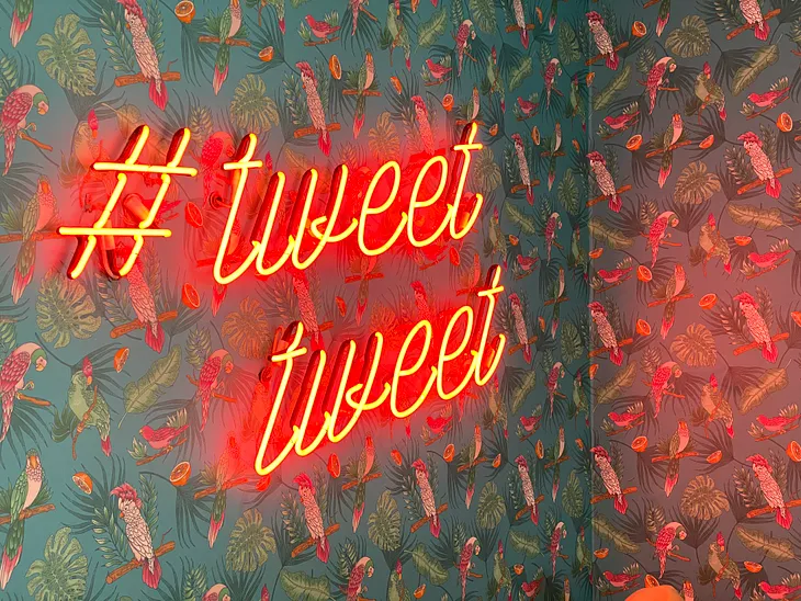 A cropped photo of the corner of a tropical inspired bird-wallpapered  room with a neon sign hung on the adjacement wall of that corner reading “#tweet tweet”; second one on top of the other.