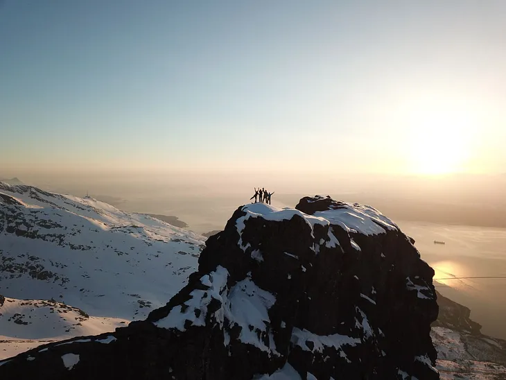 Four people standing on a rocky mountain topped with snow, more rocky peaks and the sunset in the background