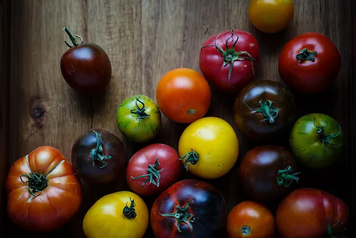 A rainbow of various sized tomatoes with most of them with green leaves and stems attached, placed on a wooden board.