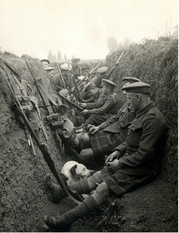 A black and white photograph of soldiers sitting silently in a trench and wearing kilts, army coats, caps, long socks and lace-up boots. Rifles and kitbags rest on the trench walls and some are held by soldiers, while a dog sits near a soldier at the front of the picture.