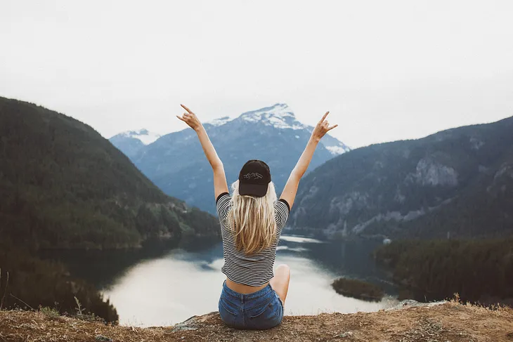 Woman with her hands outstretched high, sitting back to camera, looking towards view of mountains and lake.