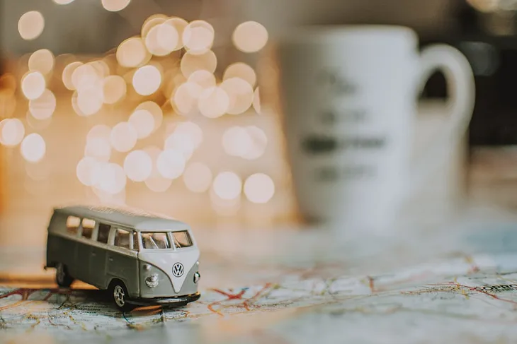 a toy VW campervan on a map on a table, in the background, out of focus, are fairy lights and a mug.