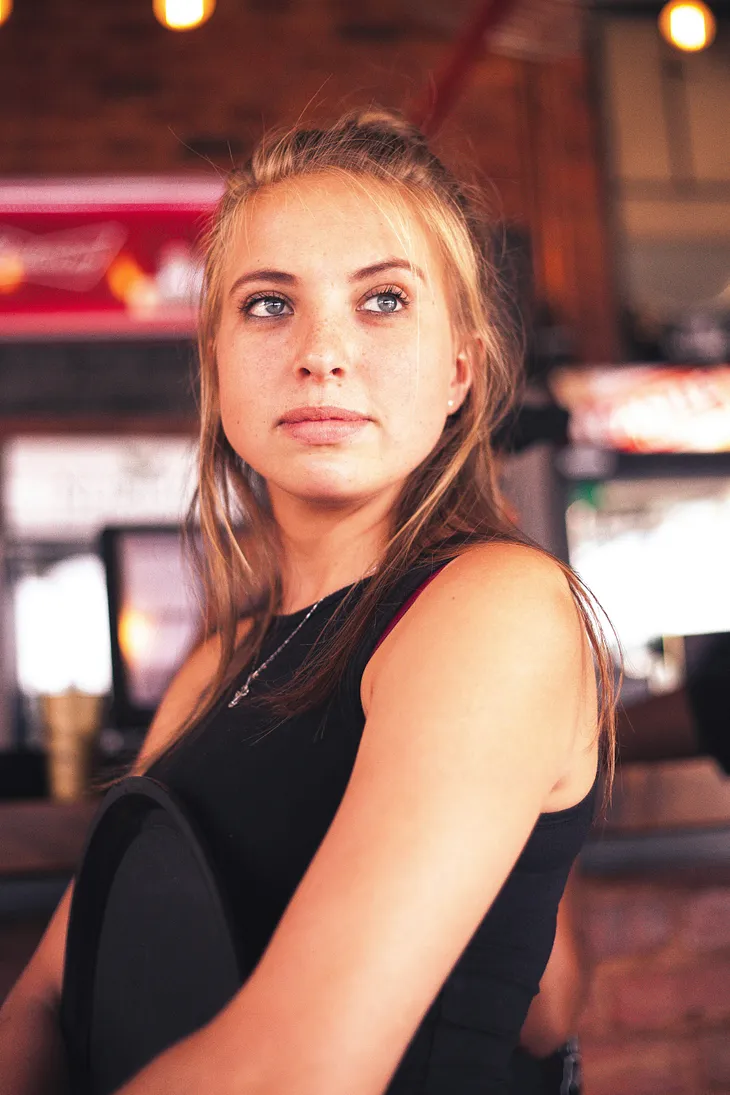 A young server with long blonde hair and wearing a black dress looks towards the camera as if in deep thought