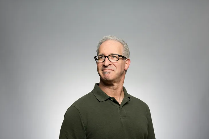 Grey-haired man with glasses, dark shirt, against a plain grey background, looking like he has synaesthesia and somebody’s just said a certain S-word.