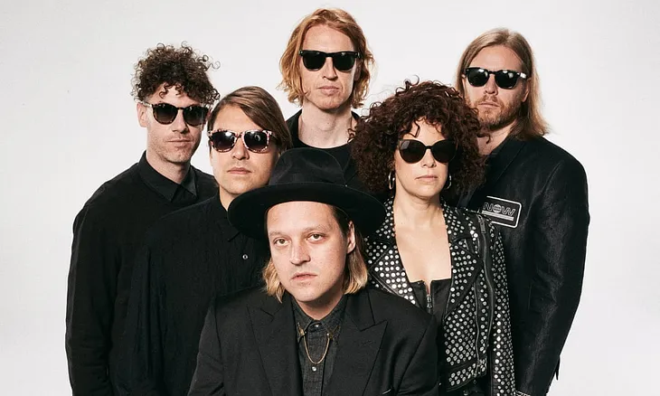 Arcade Fire band photo; all band members in black and everyone but frontman at bottom center in black sunglasses (instead wearing a black fedora) on a greyed-white background