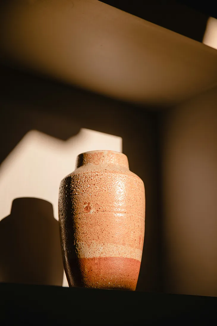 A solo urn sitting on a shelf with daylight cast upon it.