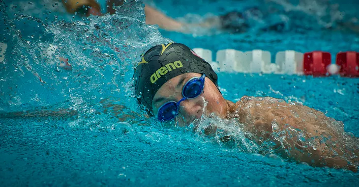 Swimmer in pool, on side, wearing a black swim cap and blue goggles.