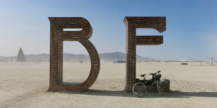 Dusty art sculpture in the open desert at Burning Man, made of light-rust color metal, spelling the word “BE” over eight feet tall.