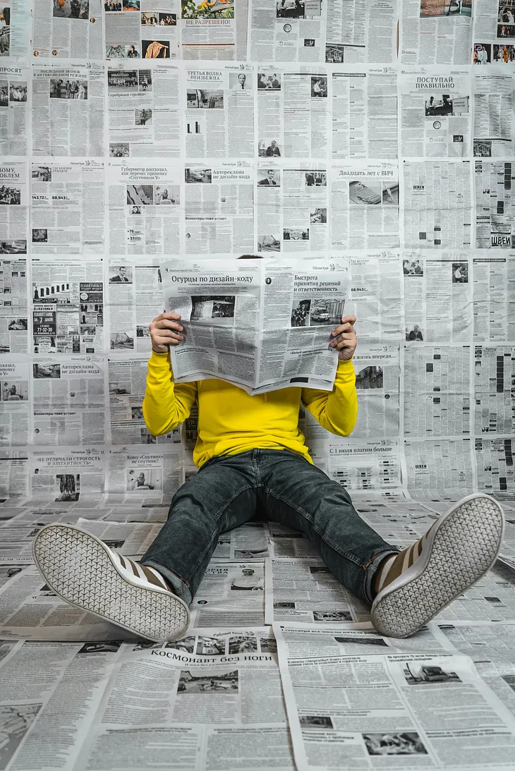 A man in a yellow shirt is reading the newspaper. He’s sitting on the ground in a room that is papered with newsprint.