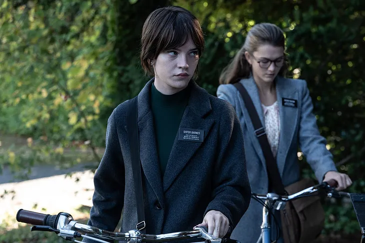 Two female Mormon missionaries, looking miserable, push their bikes along a leafy street.