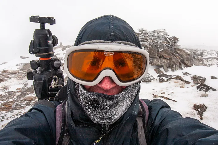 A selfie of a frost-covered man with a tripod, a frozen summit cairn in the background.