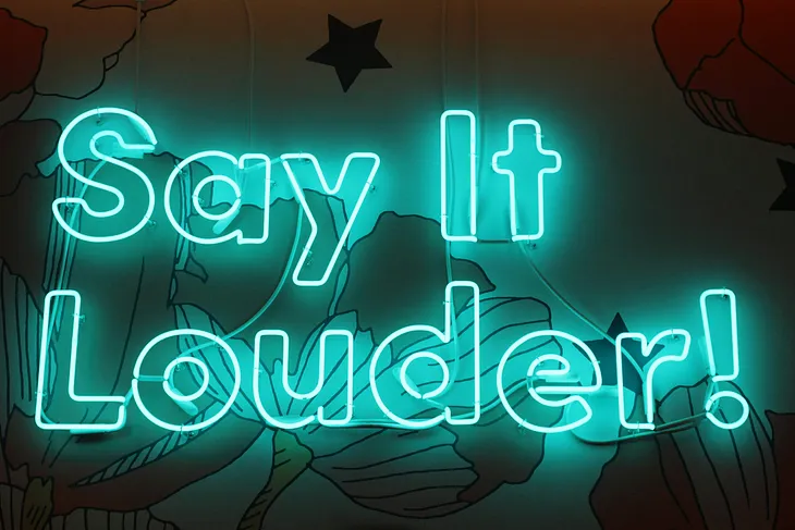 Neon sign that reads “Say it louder!”