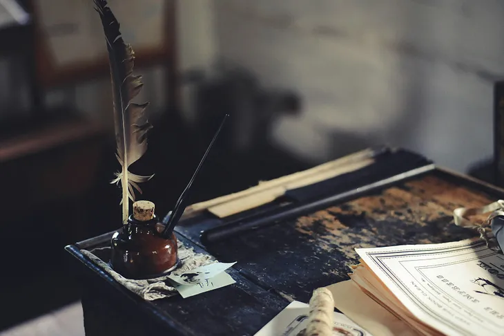 Vintage feather pen & calligraphy pen with ink bottle at the corner, and sheets of script on desk.