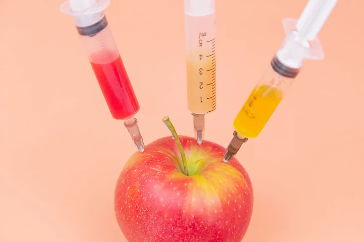 Three syringes stick out of an apple. Each syringe is filled with a different colored liquid. One is red, one is yellow, and the other is orange.