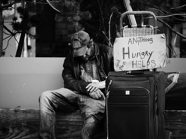 A dejected looking  man sitting with a suitcase and a sign lettered “Hungry Anything Helps” and “God Bless”