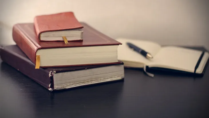 A stack of three hardcover books lies on a dark surface, with the top book open slightly to reveal a ribbon bookmark. Next to the stack, a pen rests on an open notebook with blank pages