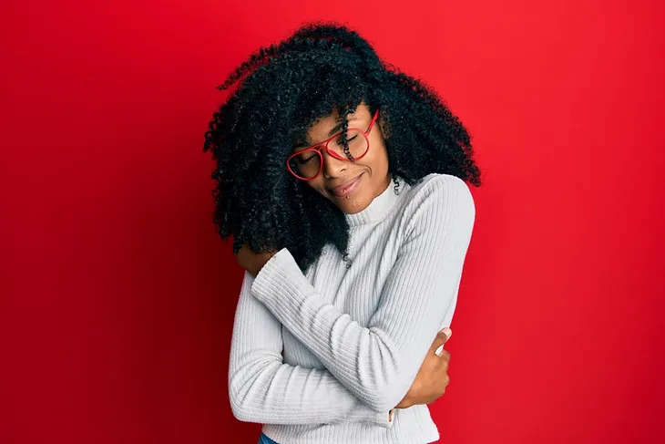 A photo of a black person wearing a white turtleneck shirt and red glasses. Their eyes are closed and they are smiling as they hug themself.