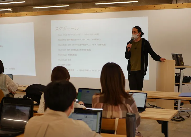 Tetsu, a Japanese man, stands in front of a whiteboard with a projection with an audience of students on their computers looking up at him.