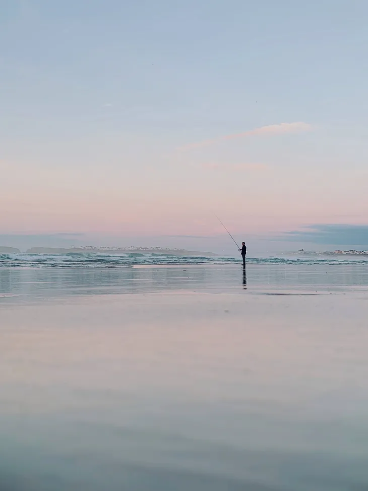 A lone fisherman on a calm beach facing the waves.