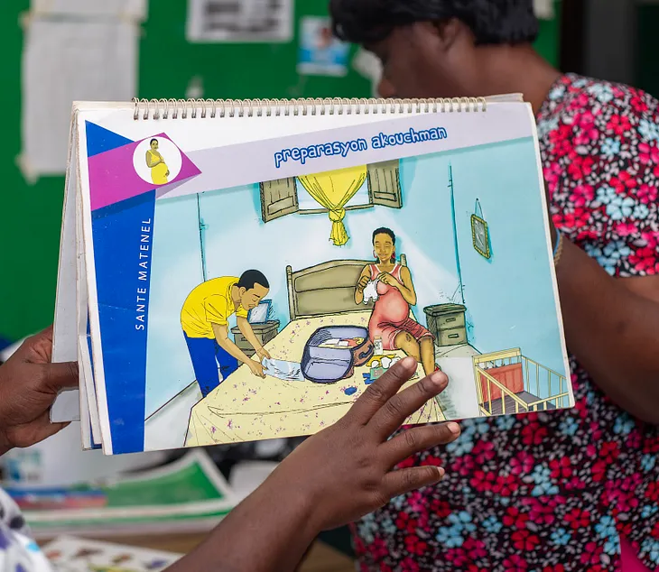 A person is holding a training aid that shows a pregnant mother sitting on a bed with her husband on the other side, prepping for the arrival of their child.