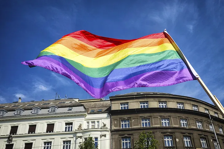 A large rainbow Pride flag waves in the wind against a bright blue sky, above a row of historic European-style buildings.