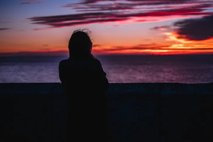 Silhouette of woman looking longingly out to sea at sunset. The sunset is super-duper pretty.