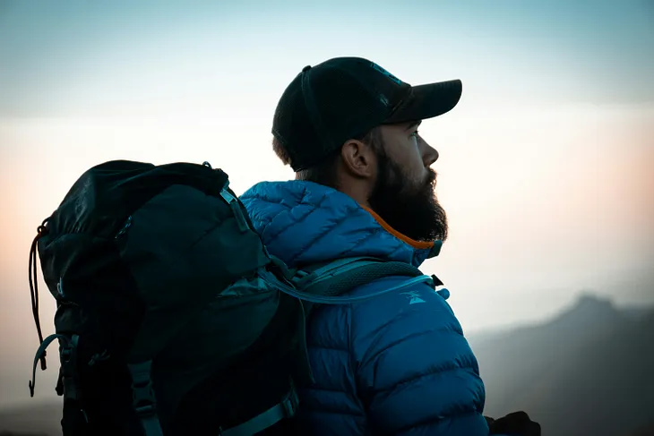 A young, bearded hiker with backpack stares into the distance